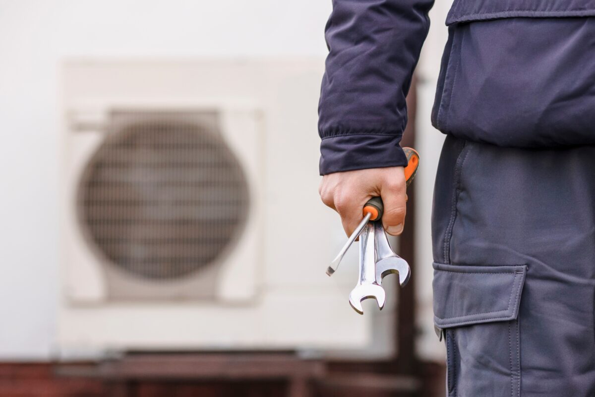 Worker hand holding tools with a heat pump in the background.