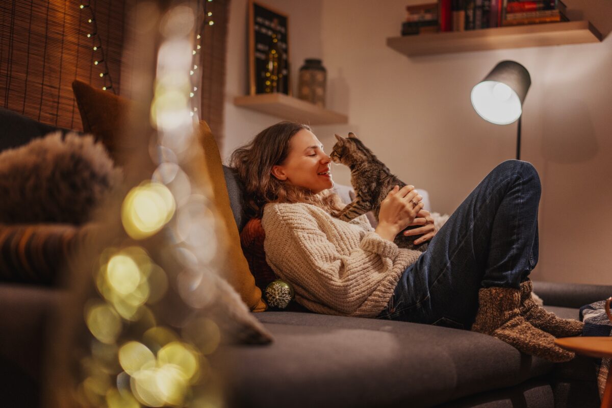 Woman relaxing on a sofa at home in the evening with a tabby cat beside her.