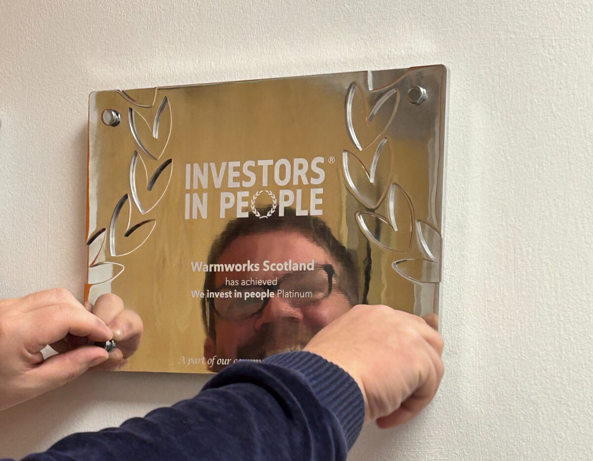 A man smiling, hanging a plaque that reads: "Investors in People. Warmworks Scotland has achieved We invest in people Platinum."